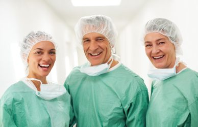 Trije zdravniki v zelenih operacijskih oblekah in kapah na bolnišničnem hodniku